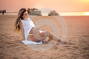 Beautiful young pregnant woman sitting on sand, touching her belly and enjoying pregnancy on the beach at sunset.