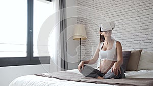 Beautiful young pregnant woman using virtual reality glasses while sitting on the bed at home