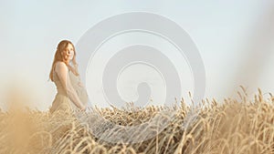 Beautiful young pregnant woman walks on wheat field at sunset, expectant mother with relax in nature stroking her belly with hand