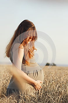 Beautiful young pregnant woman walks on wheat field at sunset, expectant mother with relax in nature stroking her belly with hand