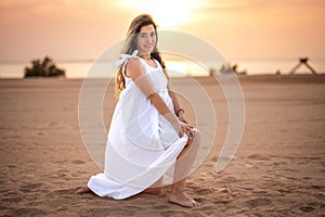 Beautiful young pregnant woman in white dress posing on the beach at sunset.