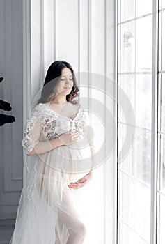beautiful young pregnant woman in white dress stands near window