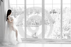 beautiful young pregnant woman in white dress stands near the window black white photo