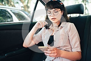 Beautiful young pretty girl is using a smart phone and smiling while sitting on back seat in the car