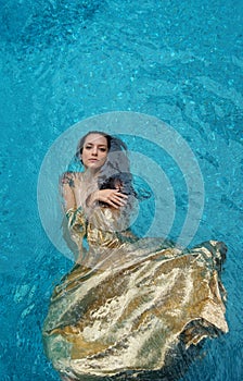 Beautiful young proud woman in golden dress, evening dress floating weightlessly elegant floating in the water in the pool dark