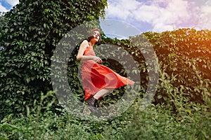 Beautiful young rebel brunet girl in red dress in green park bushes walking in black shoes