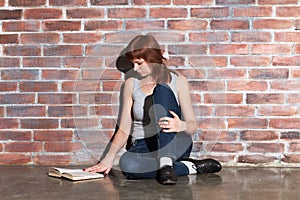 Beautiful young red hair woman in jeans with book sitting on floor near brick wall. Attentively reading an interesting