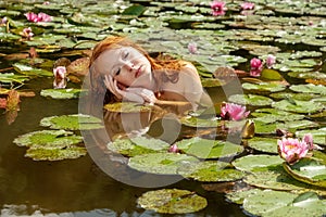 Beautiful young red-haired mermaid woman sensually seductively delights in the water, with pink water lilies and rests on her