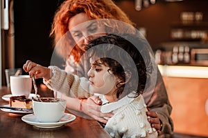 Beautiful young red-haired mother with cute curly-haired daughter are sitting in cozy cafe and drinking hot school. Mothers Day.