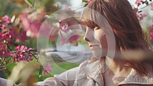 Beautiful young red-haired woman among cherry blossom sakura tree spring pink flowers making selfie on smartphone on sunset