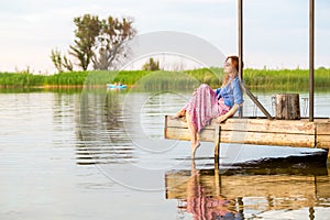 Beautiful young red woman sitting on a wooden pier in a colorful long sarafan dress and putting her leg in a water