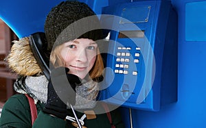 Beautiful young redhead girl in cold season outfit using street blue payphone.