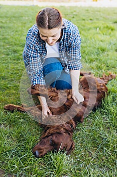 Beautiful young redhead woman playing with happy irish Setter dog in a park outdoors. Girl sitting on a green grass with
