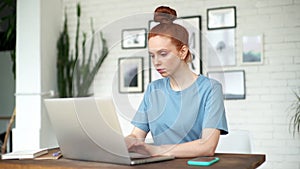 Beautiful young redhead woman is working on laptop at the desk in room at home.