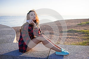 Beautiful young redheaded woman sitting on waterfront, enjoying nature and the good weather. The girl in red plaid shirt