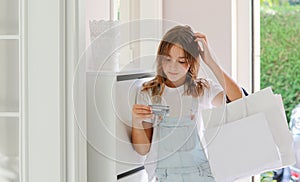 Beautiful young reflective teenager girl with shopping bags looking at credit card scratching her head.