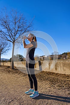 Beautiful young runner girl, beautifully illuminated by sunrise light, drinking water and hydrating from a bottle, dressed in