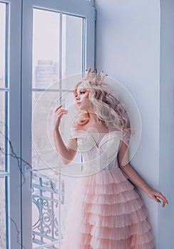 Beautiful young sad fairy tale cute queen woman stand near vintage antique window. Girl Princess blond long wavy hair