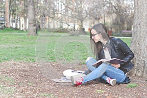 Beautiful young school or college girl with long hair, eyeglasses and black leather jacket sitting on the ground in the park readi