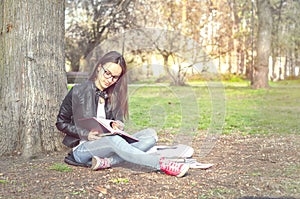 Beautiful young school or college girl with long hair, glasses and black leather jacket sitting on the ground in the park reading