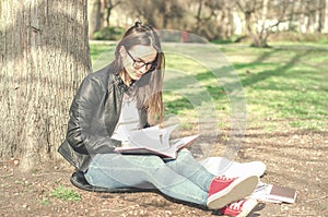 Beautiful young school or college girl with long hair, glasses and black leather jacket sitting on the ground in the park reading