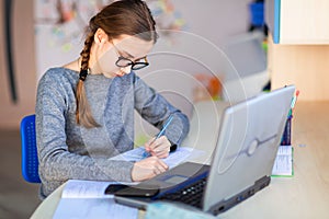 Beautiful young school girl left-handed working at home in her room with a laptop and class notes studying in a virtual class.