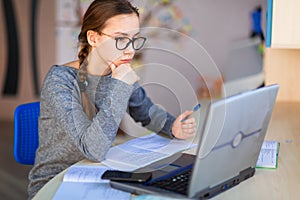 Beautiful young school girl left-handed working at home in her room with a laptop and class notes studying in a virtual class.