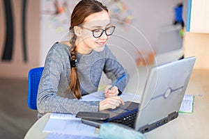 Beautiful young school girl left-handed working at home in her room with a laptop and class notes studying in a virtual class.