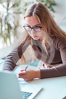 Beautiful young school girl left-handed working at home in her room with a laptop and class notes studying in a virtual class.