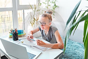 Beautiful young school girl left-handed working at home in her room with a laptop and class notes studying in a virtual class.