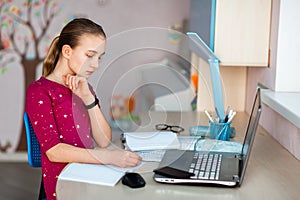 Beautiful young school girl working at home in her room with a laptop and class notes studying in a virtual class. Distance