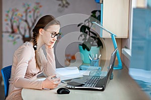 Beautiful young school girl working at home in her room with a laptop and class notes studying in a virtual class. Distance