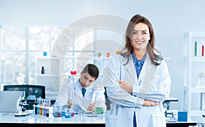 A beautiful young scientist stands smiling in a science lab with a male scientist sitting in the back