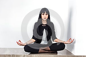 Beautiful young serious woman dressed in a black business suit sitting in lotus position on a floor in office, looking to camera