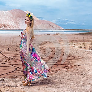 Beautiful young girl model with long red hair in a beautiful wreath of flowers and a long bright colored dress in the desert