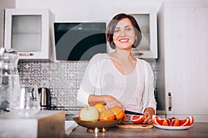 Beautiful young sincerely smiling female chopping grapefruit using a knife and cutting board in modern kitchen. Plenty of apples,