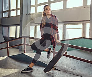 A beautiful young skateboarder girl wearing a checkered shirt sitting on a grind rail in skatepark indoors.