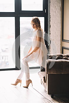 Beautiful young slender blond woman with curly hair in white trousers, a blouse and shoes in a loft studio near the window. Soft