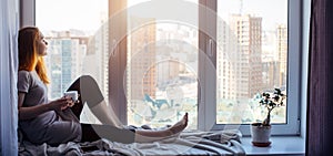 Beautiful young slender girl sitting on the windowsill at home, side view, copy space. Outside the window sky and tall city