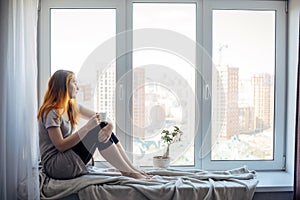 Beautiful young slender girl sitting on the windowsill at home, side view, copy space. Outside the window sky and tall city