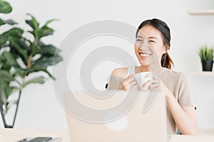 Beautiful young smiling asian woman working on laptop and drinking coffee in living room at home.