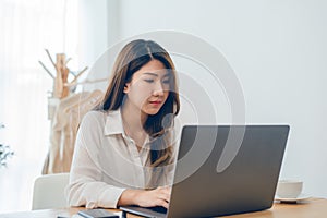 Beautiful young smiling Asian woman working on laptop while at home in office work space.