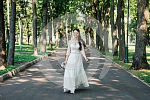 Beautiful young smiling brunette bride in wedd dress with bouquet of flowers in hands goes in the park outdoors