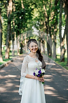 Beautiful young smiling brunette bride in wedd dress with bouquet of flowers in hands outdoors on the background of green leaves