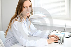 Beautiful young smiling female doctor sitting at the table near window in hospital