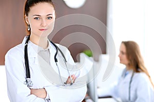 Beautiful young smiling female doctor standing in a hospital with her colleague in the background