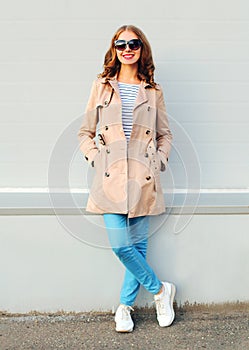 Beautiful young smiling girl wearing a beige coat and black sunglasses standing over grey