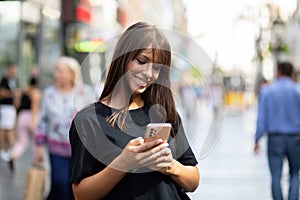 Beautiful young smiling woman in black shirt texting on smartphone on street in city center