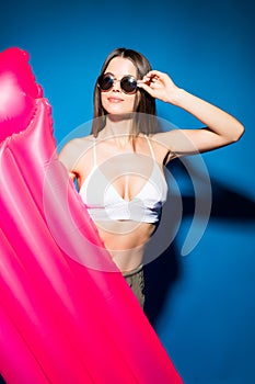 Beautiful young smiling woman dressed in white swimsuit and glasses posing with pink inflatable mattress isolated over blue backgr