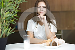 Beautiful young smiling woman eating cupcakes and croissants and drinking milk for breakfast in the kitchen looking
